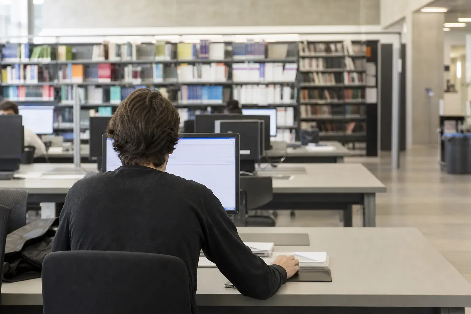 Salle de travail universitaire avec plusieurs postes informatiques et une personne de dos consultant un écran